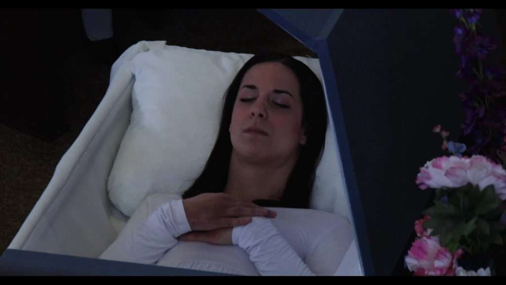Scene from movie Godforsaken. Woman in white, laying in coffin with arms crossed.